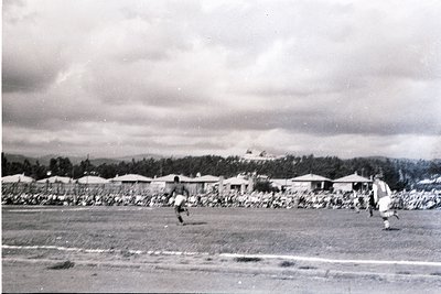Black-and-white photo of two men playing soccer on a grassy field, likely mid-20th century. Surrounding low-slung buildings w...