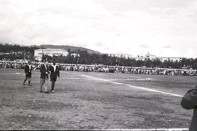 Black-and-white photo of a large-scale outdoor event on a grassy stadium field, likely a sports or public gathering. Foregrou...