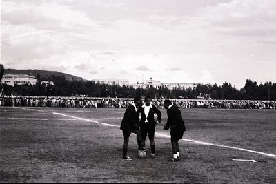 Black-and-white photo of three soccer players in vintage uniforms on a grass field, mid-game. Crowd of spectators fills stand...