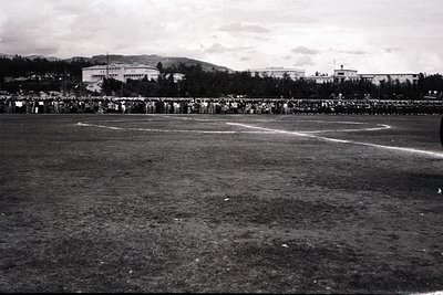 Vintage black-and-white photo of a large stadium with sparse grass field, likely for athletics or football. Crowd of spectato...