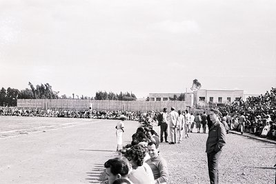Black-and-white photo of a large outdoor gathering, likely a public event or ceremony, in a courtyard surrounded by low build...