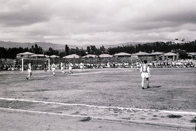 Black-and-white shot of a mid-20th-century volleyball match on a grass court, with players in knee-length shorts and knee soc...