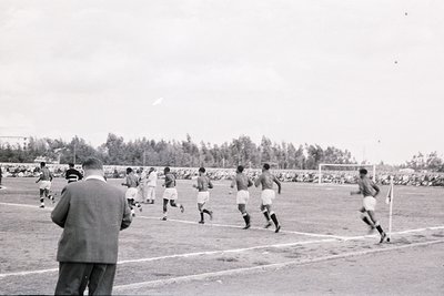 Black-and-white soccer match in mid-20th century, featuring players in white kits and dark shorts. Refereeing official in sui...