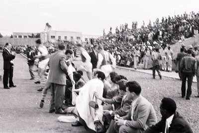 Black-and-white street scene from the 1960s–70s, featuring a crowd gathered around a central figure in a white robe. Men in s...