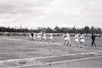 Black-and-white shot of a mid-20th-century football match, likely 1950s–60s. Players in light-colored jerseys chase a ball on...