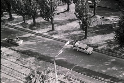 Aerial view of a mid-20th century urban street during winter, featuring a snow-covered road and sidewalk. Two vintage cars—on...