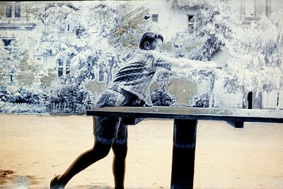 Vintage sepia-toned photo of a child mid-action, executing a table tennis forehand shot. Urban park setting with blurred gree...