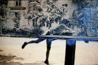 Vintage sepia-toned photo of a child mid-air on a wooden balance beam, likely mid-20th century. Urban park setting with resid...