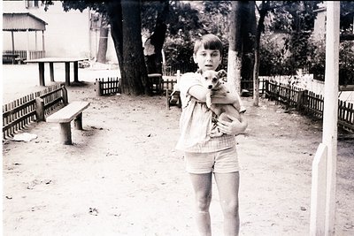 A young girl in a striped sweater and shorts holds a small dog in a park setting, likely mid-20th century. Wooden benches, a ...