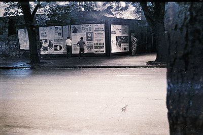 Vintage poster-covered wall in an urban setting, likely Eastern Bloc-era. Two men in light-colored shirts examine or remove p...