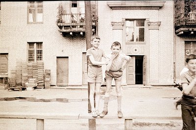 Two boys pose playfully on a low wall in an urban courtyard, mid-20th century. One boy balances on one foot while the other s...