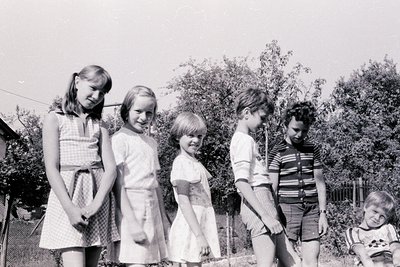 Family portrait from the 1960s–1970s, featuring five children and an adult in a backyard setting. Girls wear knee-length dres...