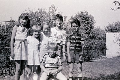 Five children pose outdoors in 1970s-era clothing: girls in knee-length dresses with pinafores, boys in striped tees and shor...