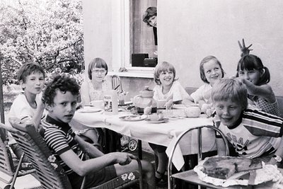 Black-and-white snapshot of seven children seated at a picnic-style table outdoors, mid-1960s. Casual summer attire—striped s...