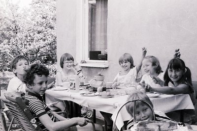 Six children seated at a long table in an outdoor dining area, mid-1960s. Lighting suggests late afternoon sun. Tableware inc...