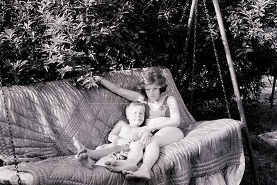 Vintage black-and-white photo of two children in a hanging chair among lush greenery, likely mid-20th century. Child on left ...