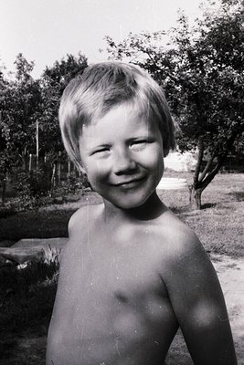 Young boy smiling outdoors, mid-20th century (likely 1950s–1970s) with blurred greenery and concrete path in background. Clas...