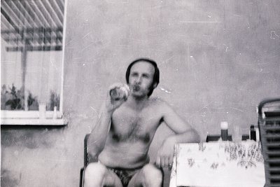 Vintage black-and-white photo of a shirtless man seated at a table, holding a cigarette in one hand and a glass in the other....