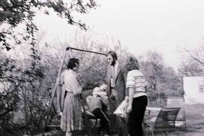 Black-and-white snapshot of three adults and a child outdoors, mid-1970s. A man in a suit and tie kneels beside a seated chil...