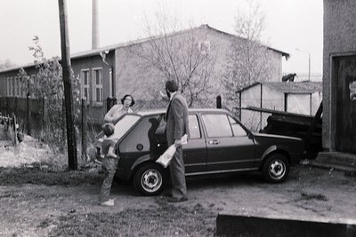 Three individuals exit a compact hatchback near a residential building in a 1970s-era urban setting. The man holds a rolled d...