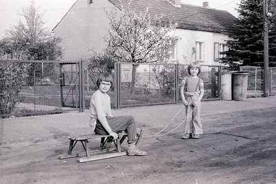 Two children play with a homemade sled in a residential courtyard, likely mid-20th century. The boy on the sled wears a short...