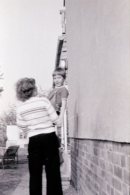 A woman in a striped sweater assists a child climbing a ladder against a brick wall, likely a residential backyard. Mid-20th ...