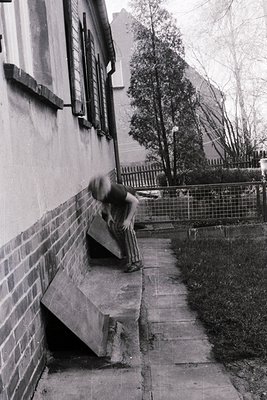 Urban street scene featuring a skateboarder mid-trick on a concrete ledge beside a brick wall. Mid-20th century residential a...