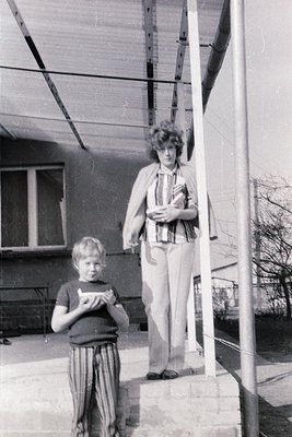 Mid-20th century black-and-white photo: woman in striped blouse and pleated skirt poses on balcony railing, holding a rolled ...