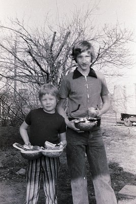 Two children pose outdoors in a rural setting, likely 1970s. Boy (left) wears striped pants and holds a woven basket with pro...
