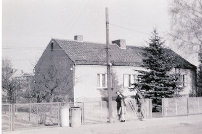 Single-story Soviet-era residential building with gable roof, concrete facade, and small rectangular windows. Two men in dark...