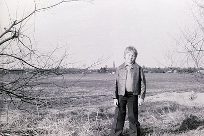 Young individual in 1970s-style jacket and trousers stands in open field with leafless trees. Rural landscape with distant ch...