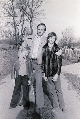 Three generations pose outdoors in rural setting, likely Eastern Europe, 1970s. Adult wears checkered shirt, wide-leg trouser...