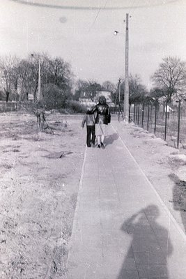 Two children walk a dog along a paved sidewalk in a mid-20th-century residential area. The paved path contrasts with unpaved ...