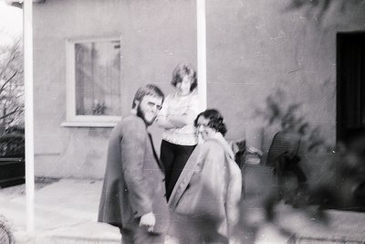 Vintage black-and-white photo of three individuals exiting a building, likely mid-1970s. Man in suit and woman in coat embrac...