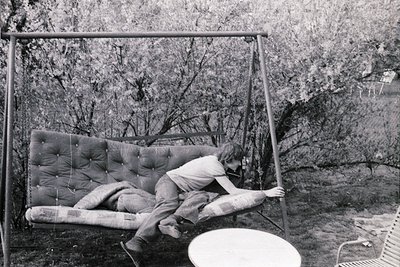 Mid-century outdoor swing suspended in a wooded area, featuring a padded seat and backrest. A person reclines on it, dressed ...