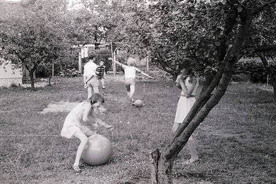 Four children play with a large exercise ball in a backyard, 1960s–70s. One kneels to push, others stand in motion. Lush gree...