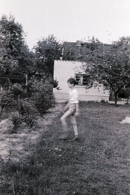 Black-and-white snapshot of a child mid-stride in a grassy backyard, 1960s-70s. Concrete path, overgrown shrubs, and a single...