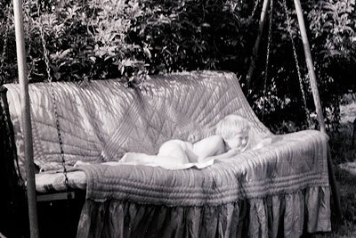 Vintage black-and-white photo of a child peacefully sleeping in a wooden swing draped with a striped blanket and ruffled hem....
