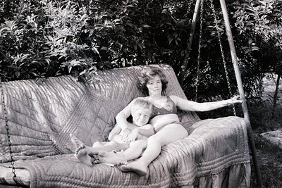 Black-and-white photo of a woman and child in a vintage hanging chair, surrounded by lush greenery. The woman holds the child...