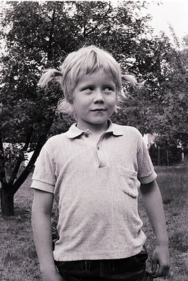 Vintage black-and-white portrait of a young boy in a short-sleeved polo shirt, standing outdoors with blurred greenery and tr...