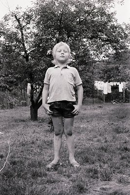 A young child stands in a grassy backyard, wearing a short-sleeved polo shirt and rolled-up shorts. Laundry hangs on a clothe...
