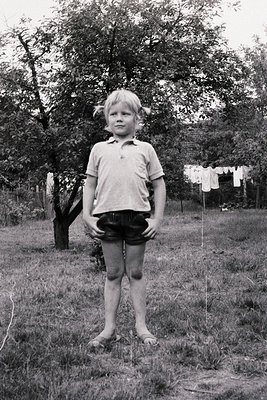 Mid-century black-and-white portrait of a young boy in a grassy backyard, wearing a short-sleeve polo shirt and rolled-up sho...