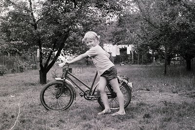 Mid-20th century black-and-white photo of a child posing on a classic bicycle in a rural backyard. The child wears shorts and...