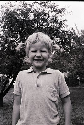 Vintage black-and-white portrait of a smiling child with light blonde hair, wearing a short-sleeve polo shirt. Outdoor settin...