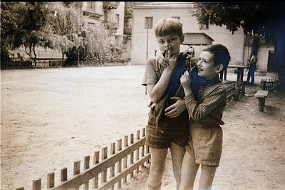 Vintage black-and-white photo of two boys in mid-1960s Eastern European courtyard. One boy holds a toy rifle, the other mimic...
