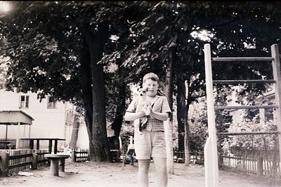 Black-and-white photo of a young boy in a plaid shirt and shorts holding a toy gun, standing in a shaded park area with picni...
