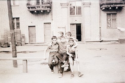 Five boys huddle in a tight group hug on a concrete ledge, set against weathered urban buildings with peeling paint and board...