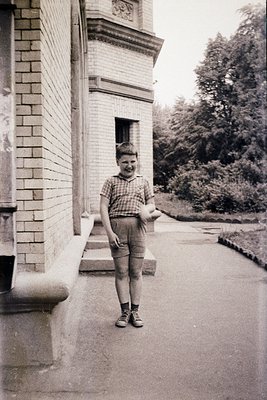 Mid-20th century boy in short-sleeved checkered shirt and rolled shorts poses outside a brick building with classical detaili...
