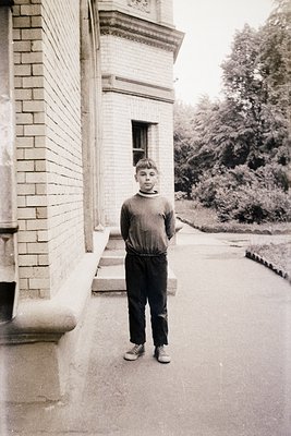 Young boy in 1970s-style sweater and baggy trousers stands beside a brick building with arched windows. Lush greenery and pav...
