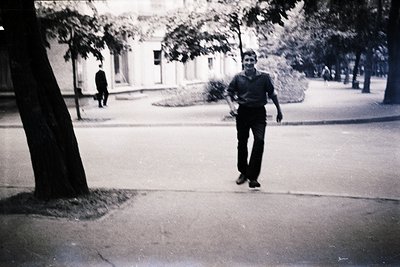 Black-and-white street scene featuring a man in a short-sleeve button-up shirt and wide-legged trousers, mid-stride on a pave...